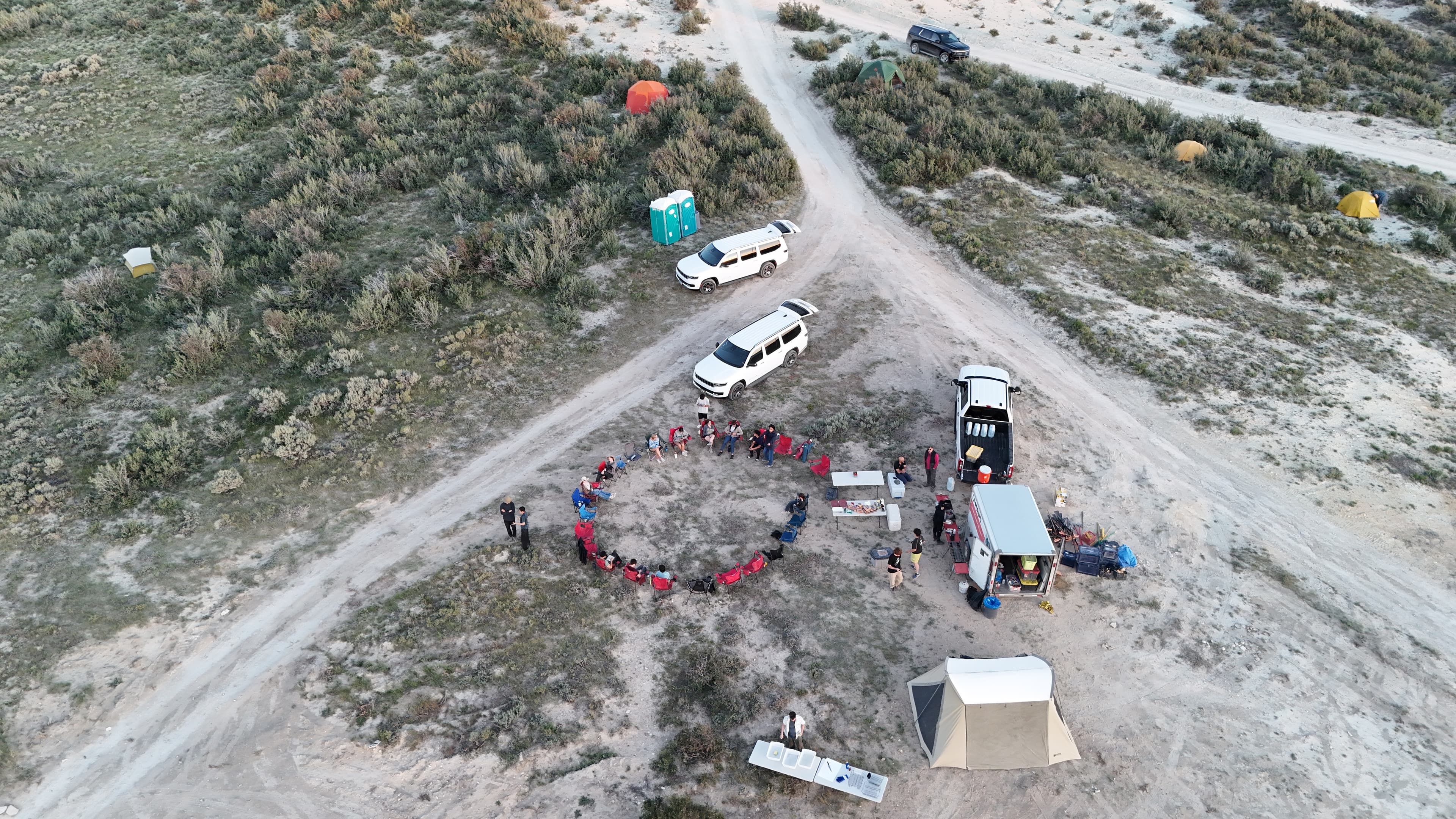 Campers gather around tables and chairs near vehicles and tents.